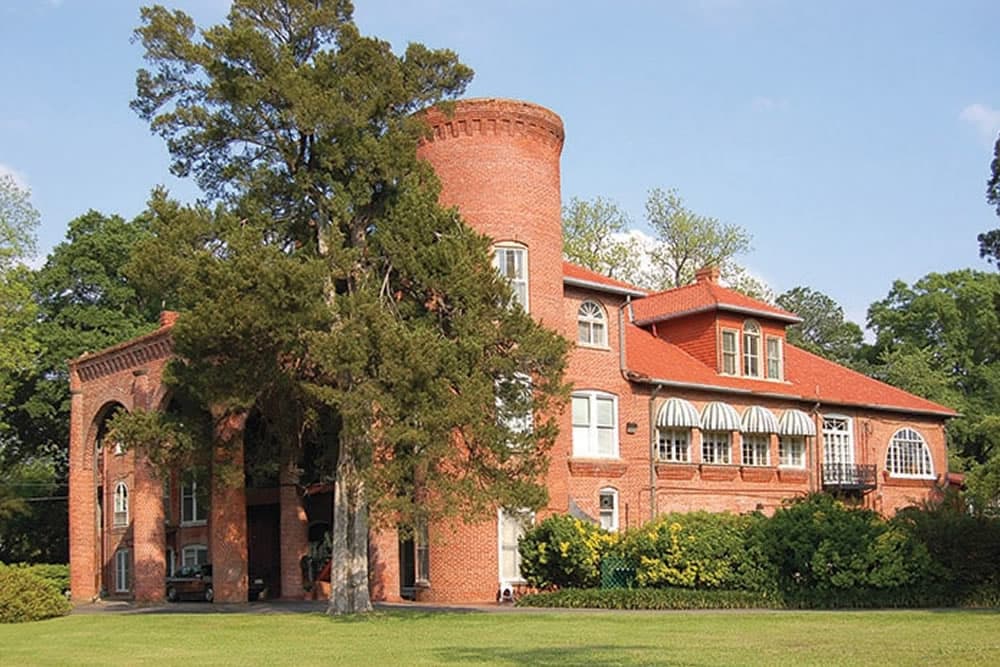 Historic red brick mansion with a tower and lush surrounding greenery.