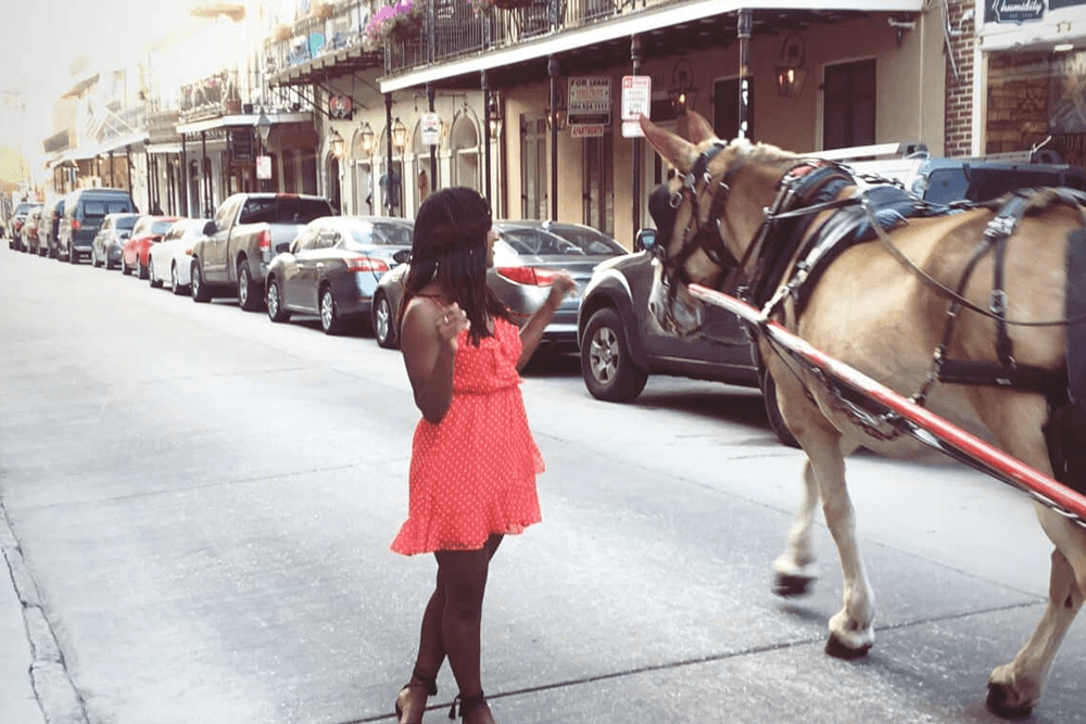 A girl in a red polka-dot dress stands on the street as a horse-drawn carriage passes by.