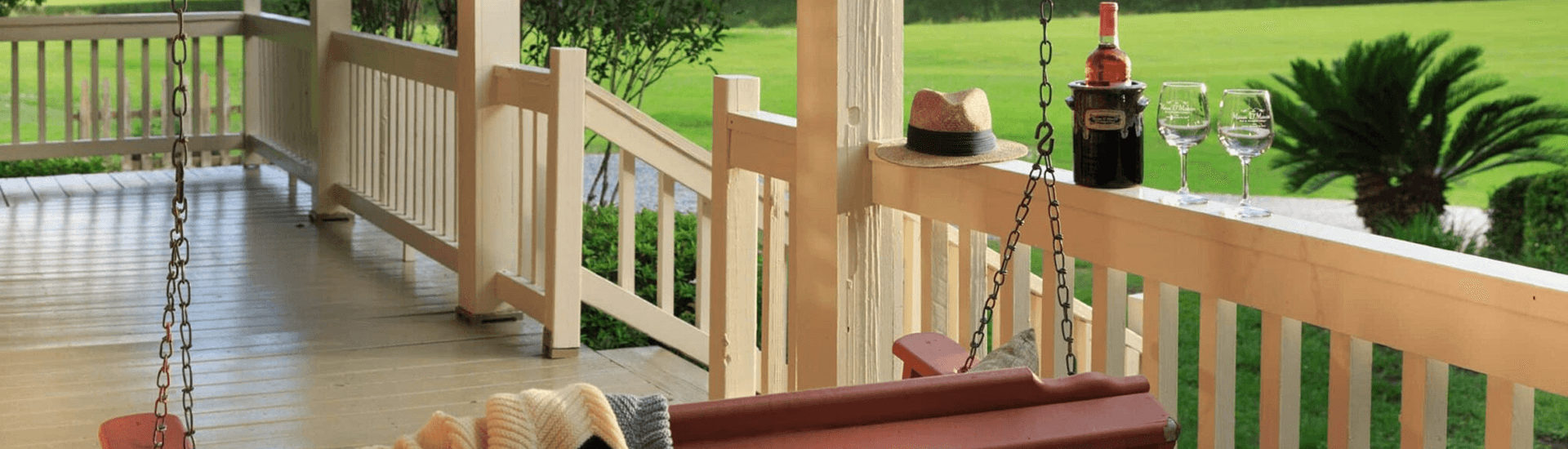 A porch swing with a cozy blanket next to a bottle of wine and glasses, overlooking a lush green lawn.